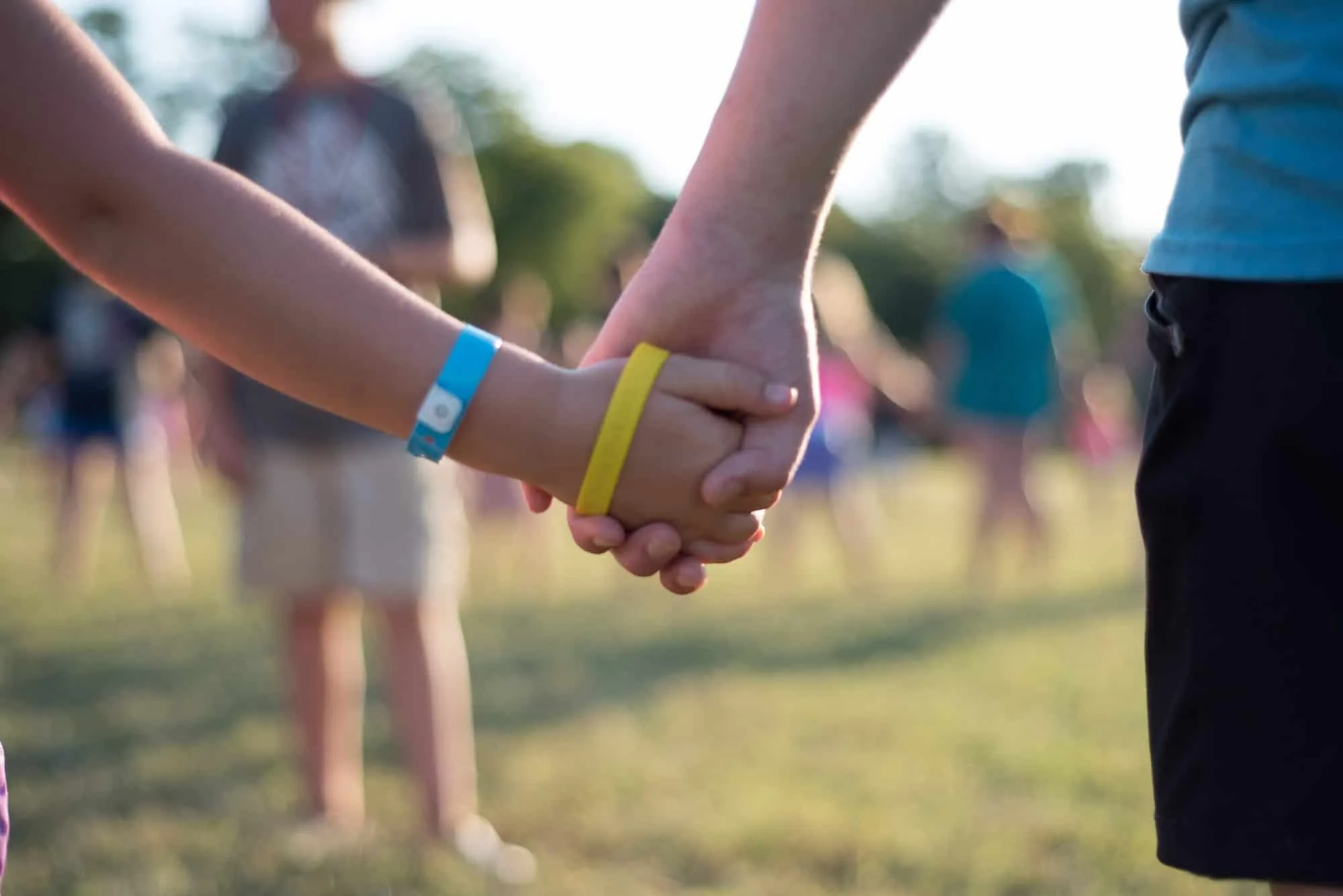 Close-up of two people holding hands outdoors. One person wears a blue wristband, and the other wears yellow bands. Blurry background shows people standing on grass in sunlight.