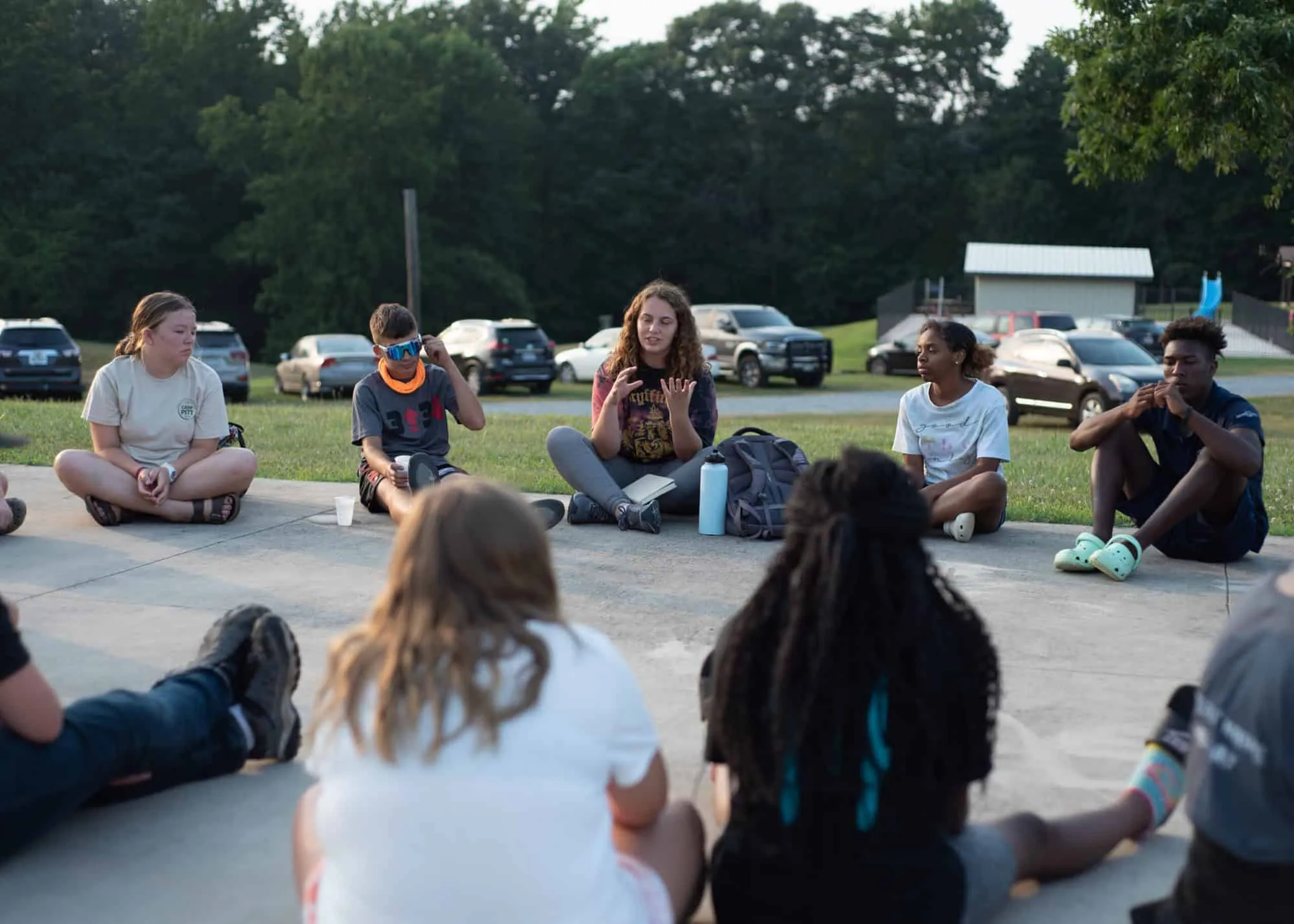 A group of young people sitting in a circle outdoors on the basketball court. They are in casual clothing, engaged in discussion, with trees and parked cars in the background.