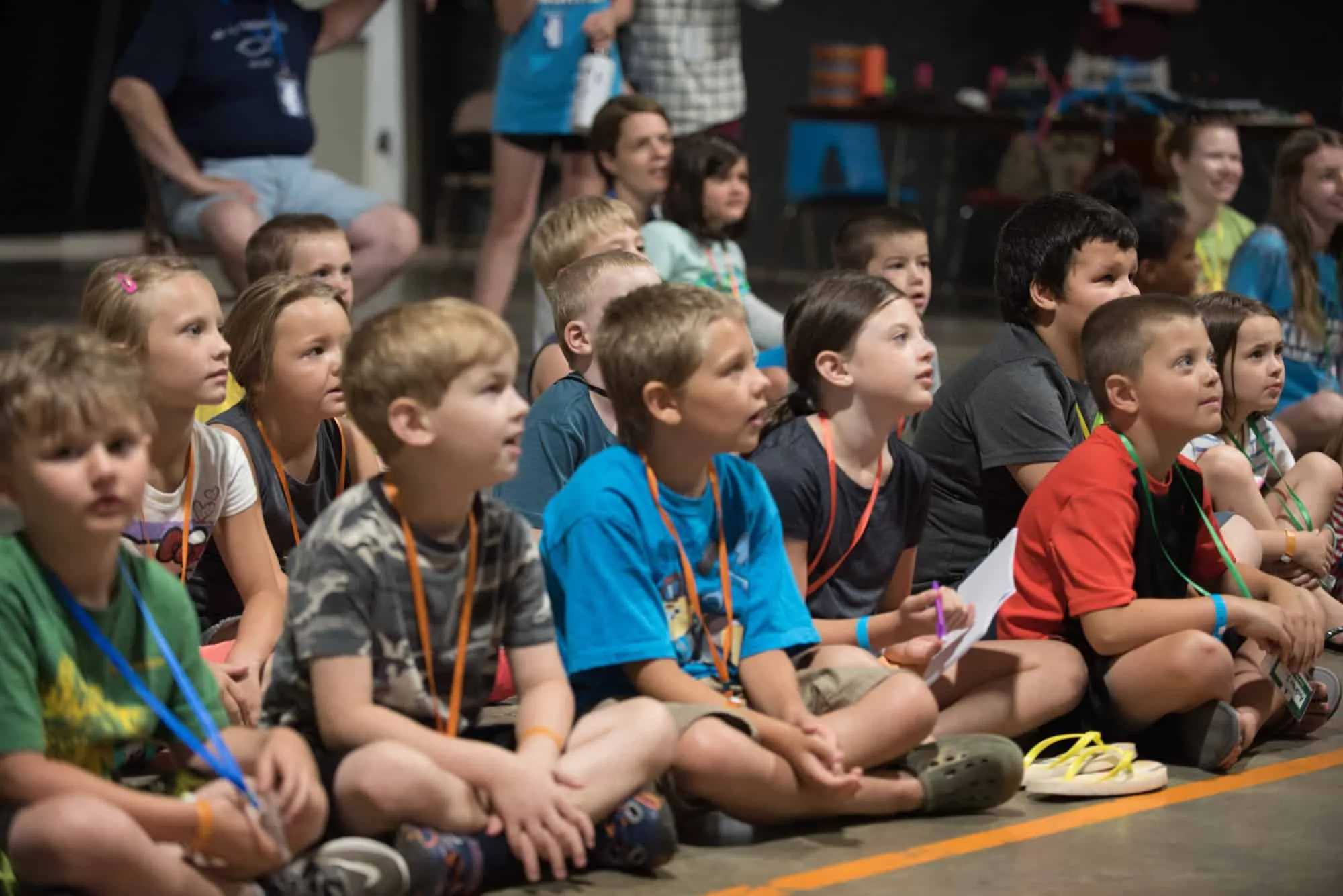 A group of young children sits on the floor, attentively watching They wear casual clothes and name tags. The background is a dimly lit indoor space, with more people seated behind them.