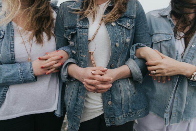 Three people dressed in denim jackets and casual tops link arms, standing closely together.