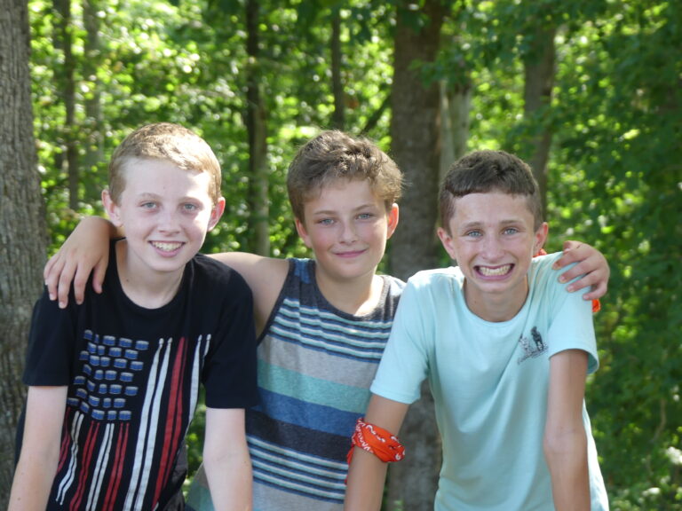 May show: Three boys stand side by side outdoors, smiling at the camera. One has an arm around the other two. Trees and greenery are visible in the background.