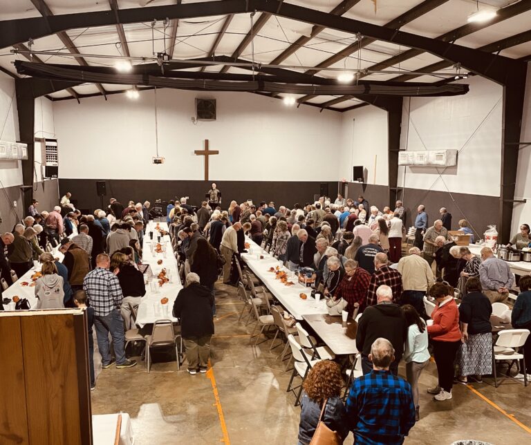 May show: A large group of people gather in a church hall with long tables for a community meal or event; a cross hangs on the back wall.