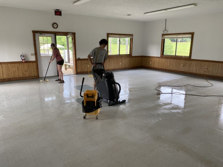 May show: Two people clean a large, empty room with white tile floors; one uses a floor scrubber while the other mops near the glass doors.