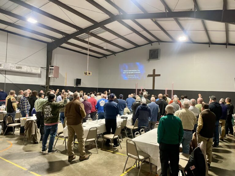 May show: A group of people stands facing a stage with a large cross and projected screen in a gymnasium, attending an indoor event with tables and chairs.