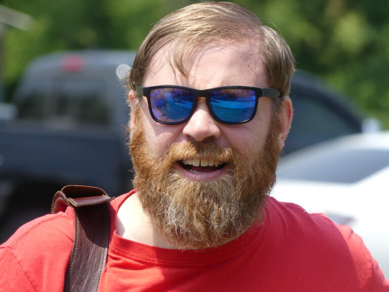May show: A bearded man wearing sunglasses and a red shirt smiles outdoors, with vehicles and greenery visible in the background.