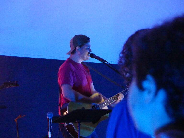May show: A musician in a red shirt and backwards cap plays guitar and sings into a microphone on stage under blue lighting. An audience member is visible in the foreground.