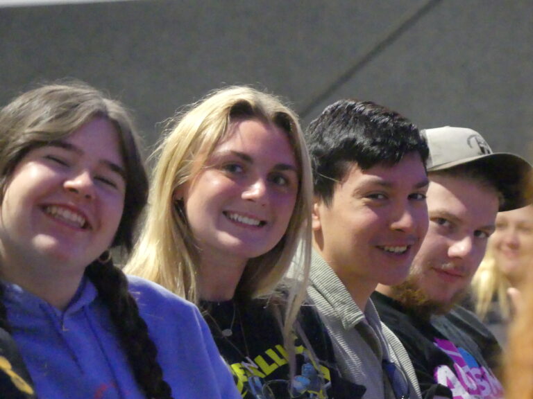 May show: Four people sitting closely together, smiling and looking toward the camera in a casual indoor setting.