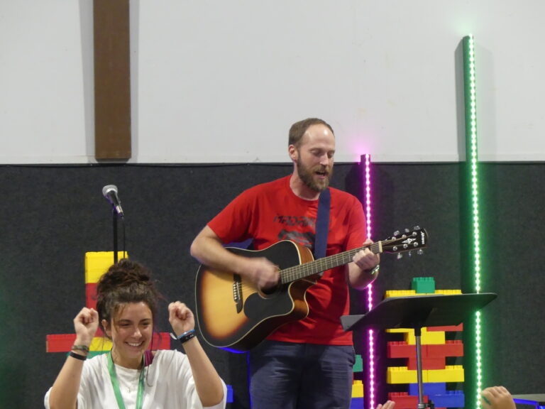 May show: A man plays an acoustic guitar and sings into a microphone while a woman in front raises her fists, with colorful blocks and LED light strips in the background.