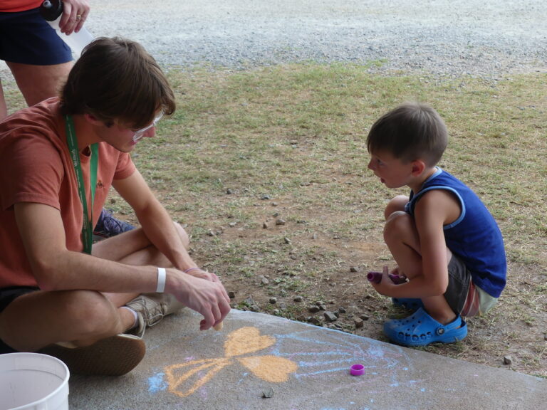 May show: An adult and a child sit on the ground outdoors, drawing with sidewalk chalk. The adult is talking to the child, who is holding a piece of chalk and looking at the drawing.