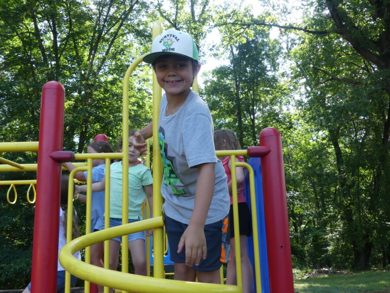 May show: A young boy in a gray shirt and white cap smiles while standing on a yellow spiral staircase at a playground, with other children playing in the background.
