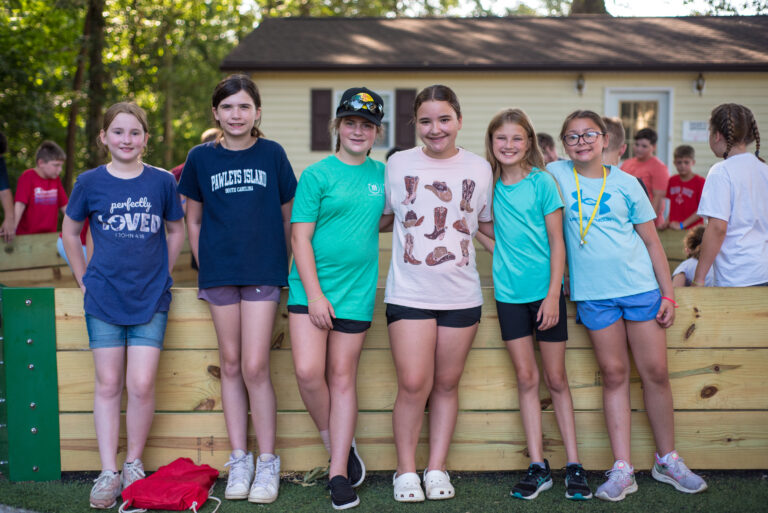 May show: Six girls stand side by side and smile in front of a wooden barrier at an outdoor camp or recreation area, with other children visible in the background.