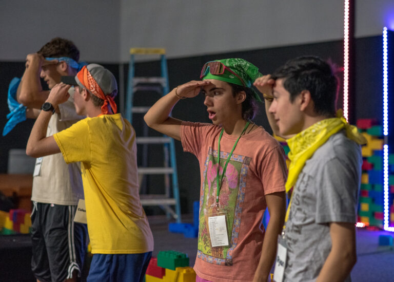 May show: Four young people wearing colorful bandanas and lanyards stand indoors, facing forward with hands raised to their foreheads as if saluting or looking into the distance.