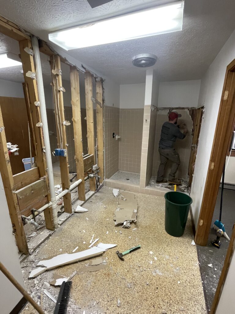 May show: A person is demolishing tiles in a partially gutted bathroom under renovation, with exposed wall studs, debris on the floor, and various tools scattered around.