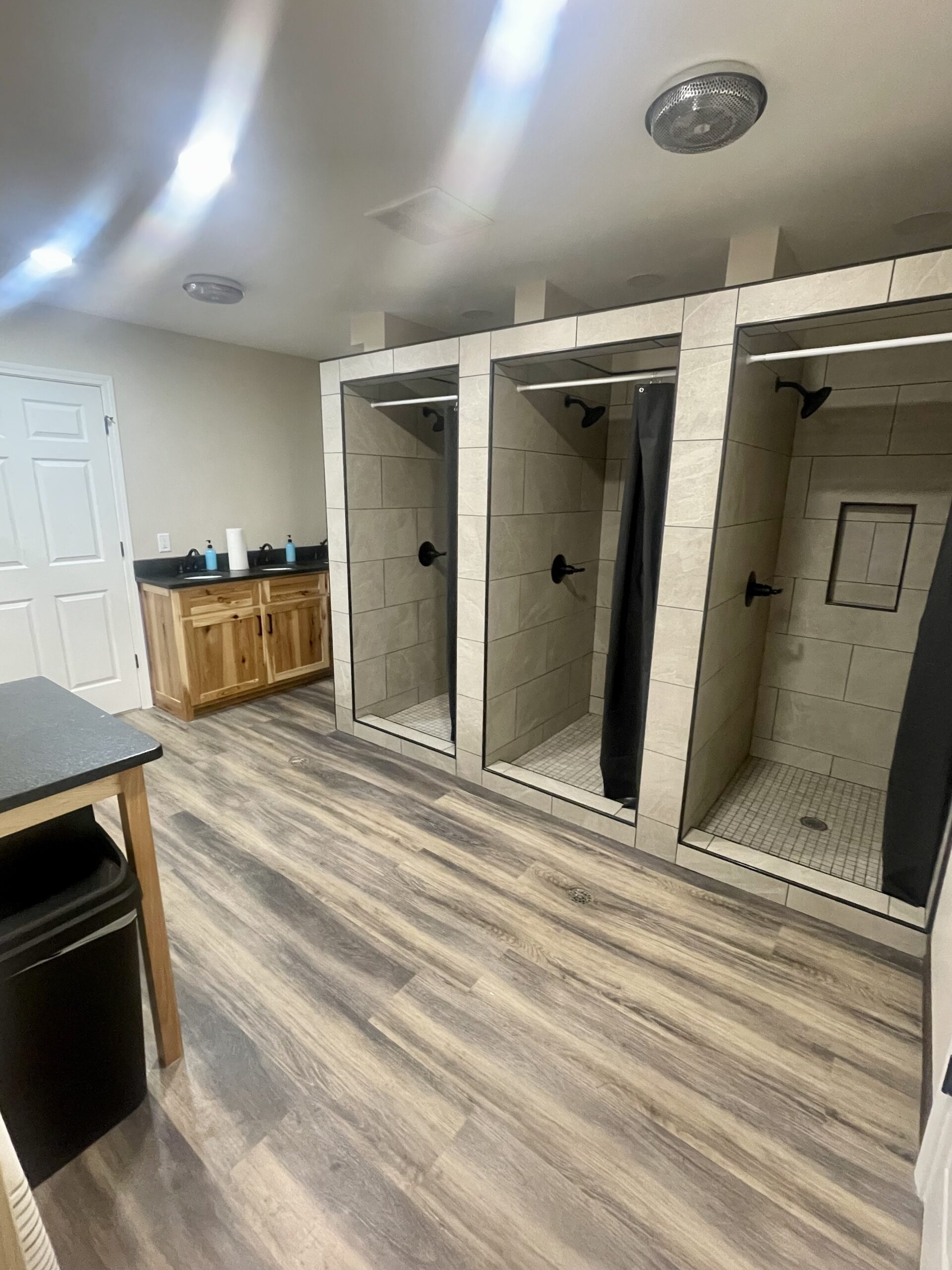 May show: A clean locker room with three tiled shower stalls, a double sink vanity, wood cabinets, and laminate wood flooring.
